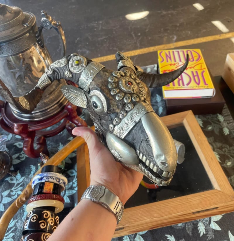A person holds an ornate metallic goat skull decorated with silver embellishments, intricate patterns, and red gemstones. Nearby are bangles, a silver teapot, framed items, and a book titled "Jack Collins.