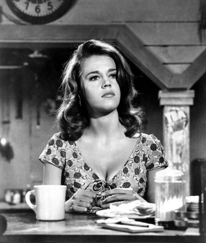 A young woman with wavy hair sits at a table in a vintage cafe, wearing a floral dress. She looks thoughtfully into the distance, with a coffee mug and a glass jar in front of her. The scene is in black and white.