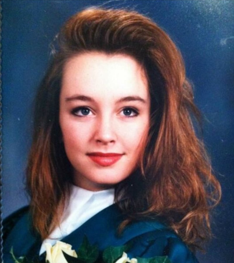 A young woman with long, wavy brown hair poses for a formal portrait in a green graduation gown with a white collar against a blue studio background. She is smiling slightly and holding a bouquet of yellow roses.