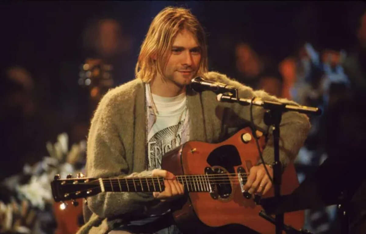 A man with long blonde hair sits on stage, wearing a gray cardigan and white t-shirt, holding an acoustic guitar and singing into a microphone in a dimly lit setting.
