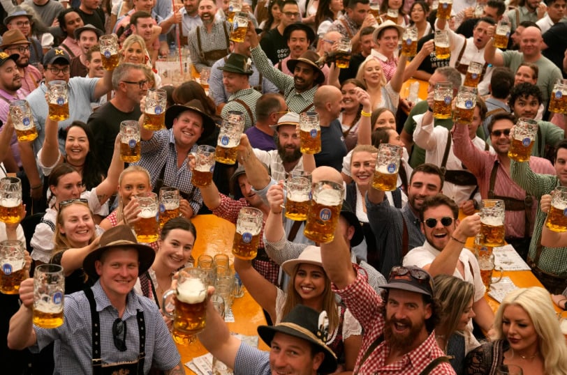 A large crowd of people dressed in festive attire smile and raise beer mugs at an outdoor event, likely a festival or celebration, with long tables and a lively atmosphere.