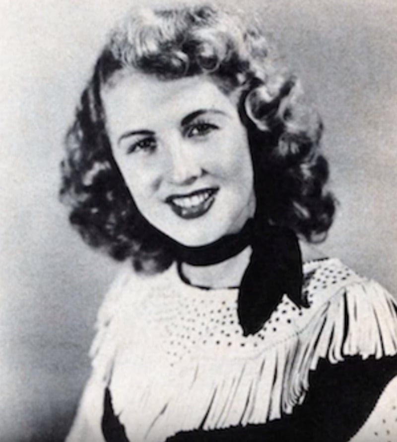 Black-and-white portrait of a smiling young woman with curly hair, wearing a fringed top and a scarf tied around her neck.