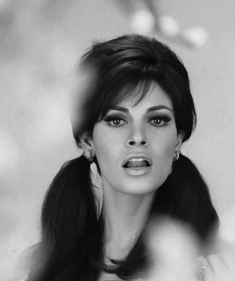 Black and white portrait of a woman with voluminous dark hair styled in two low ponytails and wispy bangs, wearing hoop earrings, looking directly at the camera with a soft, blurred foreground.