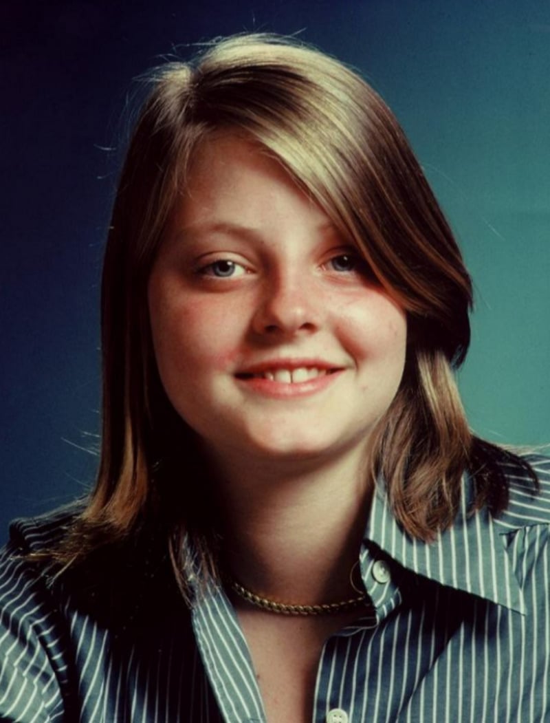 A young person with straight, shoulder-length light brown hair and fair skin smiles slightly. They are wearing a blue and white striped shirt and a gold chain necklace, against a plain blue background.