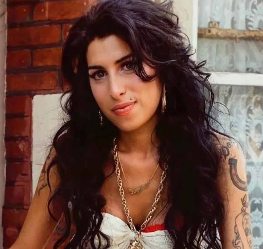 A woman with long, curly dark hair and tattoos is wearing hoop earrings, layered necklaces, and a white top. She is standing outside in front of a brick wall and a window with lace curtains, looking at the camera and smiling slightly.