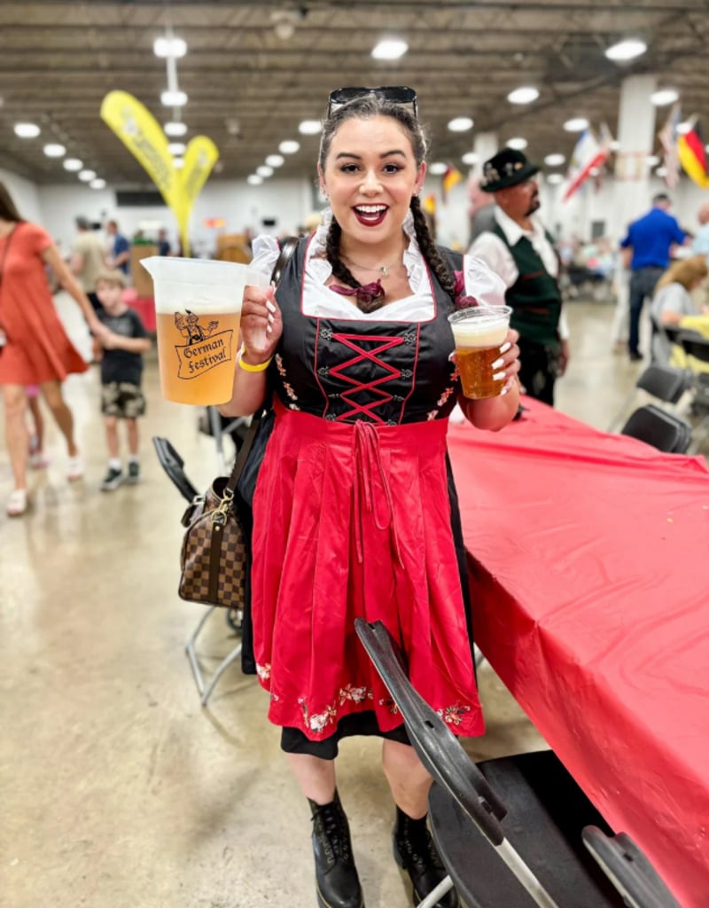 A smiling woman in a red and black dirndl dress holds a beer and a German festival stein in a lively indoor event with people, tables, and festive decorations in the background.