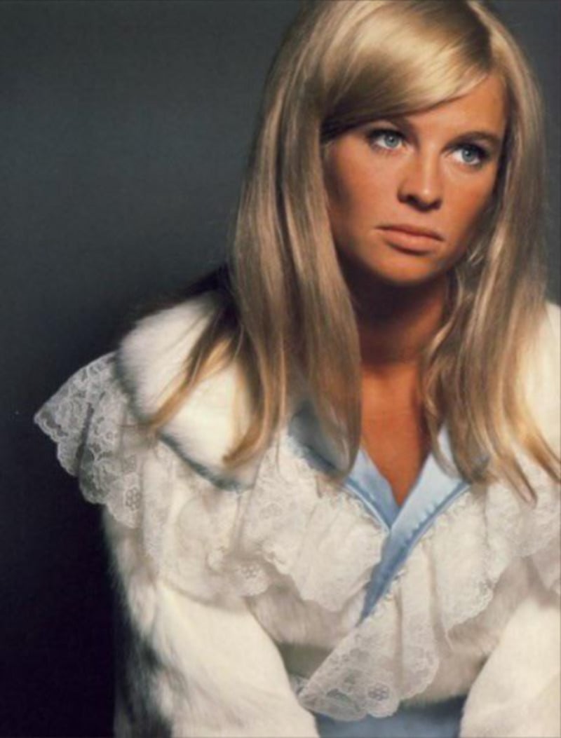 A woman with long blonde hair and blue eyes wears a white fur coat with lace details over a light blue top, looking off to the side against a plain, dark background.