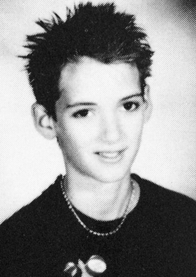 Black and white yearbook-style photo of a young person with short, spiky hair, wearing a dark shirt and layered necklaces, smiling slightly at the camera.
