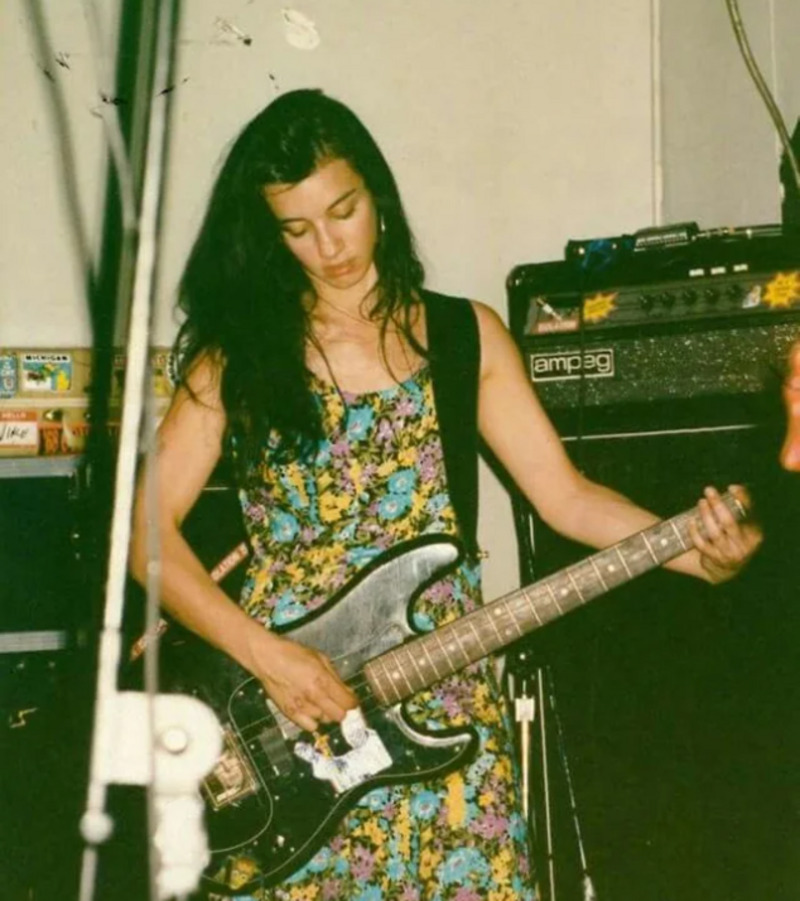 A woman with long dark hair, wearing a colorful floral dress, plays a black electric bass guitar in front of amplifiers during a live indoor performance.