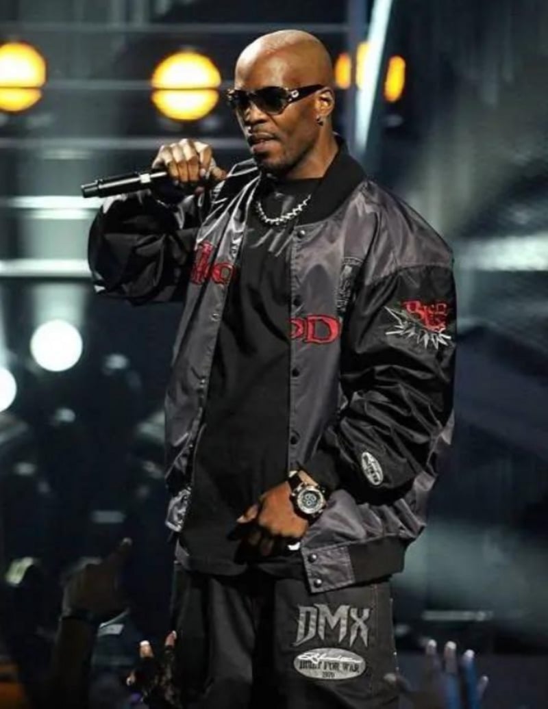 A man wearing sunglasses, a black jacket, and black pants holds a microphone on stage under bright lights, performing in front of an audience.