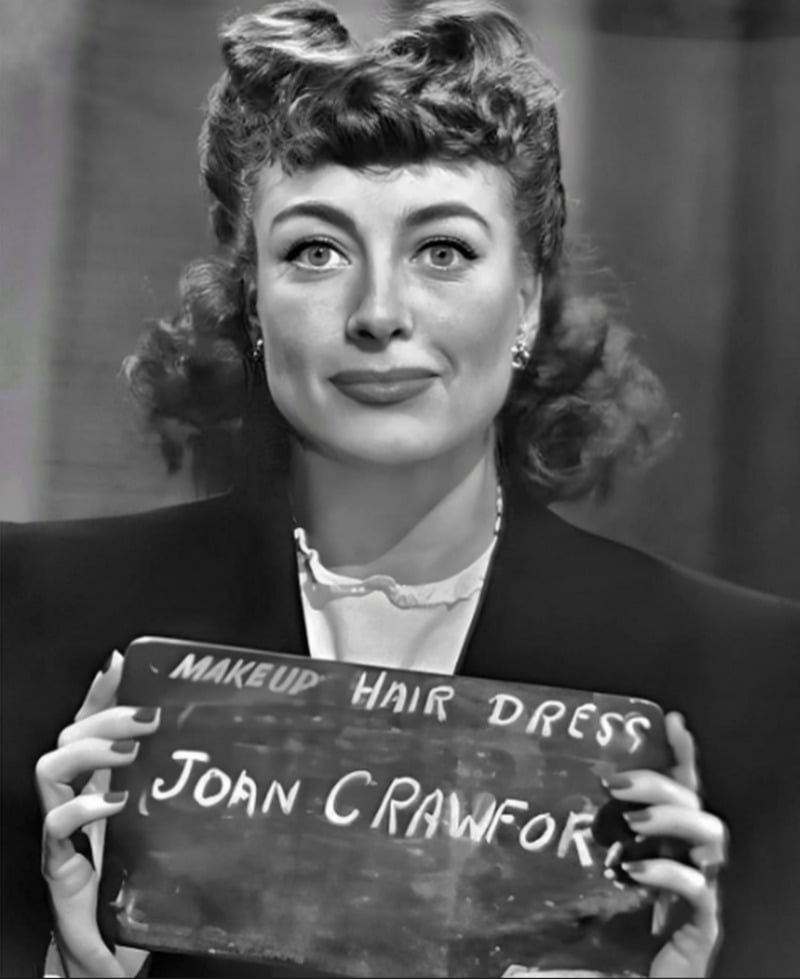 Black and white photo of a woman with wavy hair holding a sign that reads “MAKEUP HAIR DRESS JOAN CRAWFORD,” looking directly at the camera with a neutral expression.