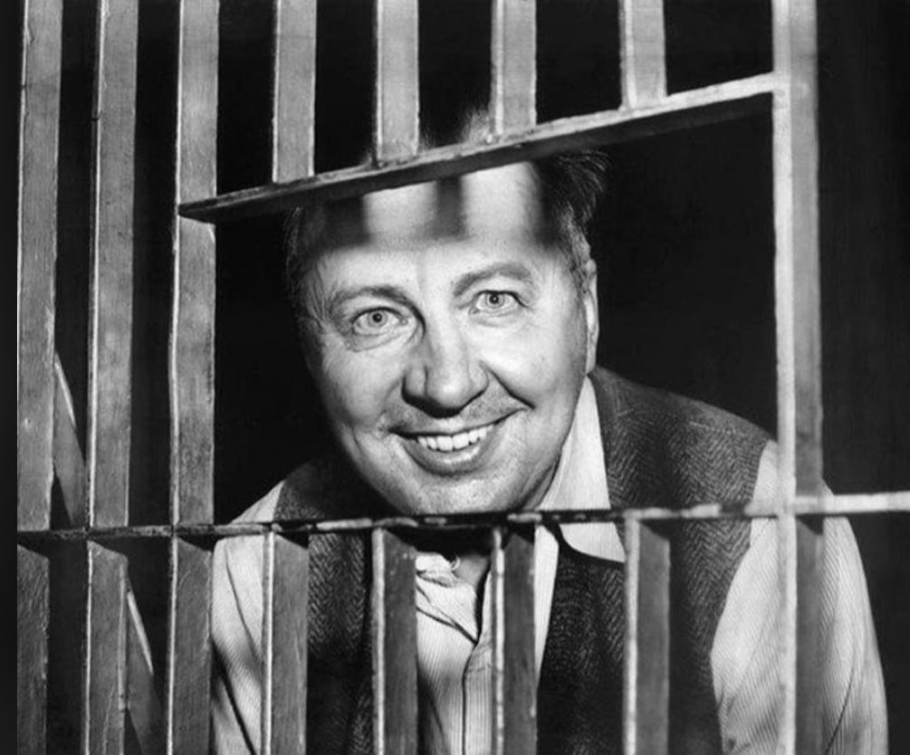 A smiling man wearing a collared shirt and vest looks through metal jail bars in a black-and-white photograph.