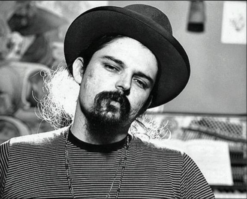 A man with long hair, a goatee, and a mustache wears a wide-brimmed hat and a striped shirt, looking directly at the camera in a black-and-white photo. The background is blurred and indistinct.