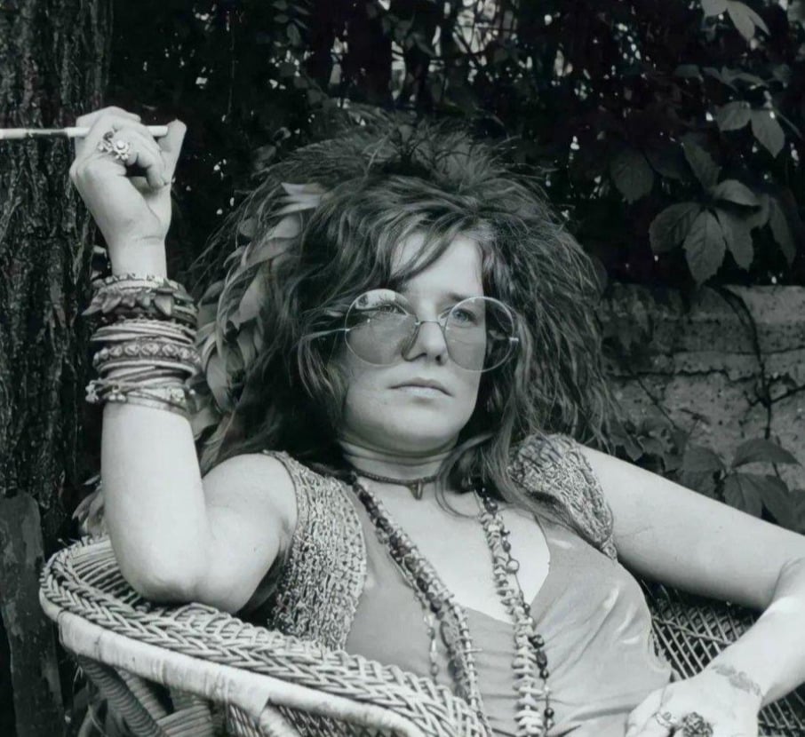 A woman with wavy hair sits in a wicker chair outdoors, wearing large round glasses, layered necklaces, and many bracelets. She holds a cigarette in one hand and gazes forward with a relaxed expression.