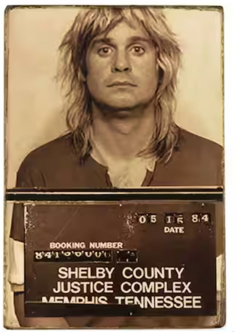 A vintage mugshot photo of a man with long blond hair wearing a two-tone shirt, holding a sign reading "Shelby County Justice Complex, Memphis, Tennessee" with a booking date of 05-18-84.