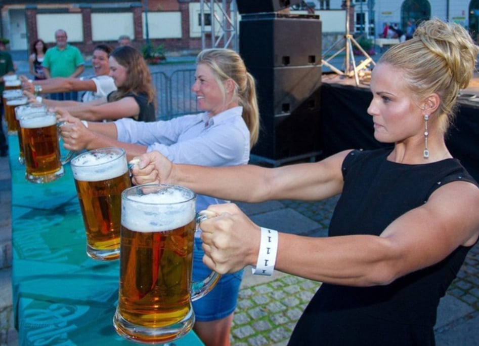 Several women compete in a beer stein holding contest, each holding a large mug of beer at arm’s length in front of them, standing outdoors with people watching in the background.