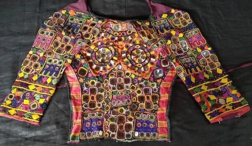 A colorful traditional blouse with intricate embroidery, mirror work, and geometric patterns in red, yellow, purple, and green, displayed on a dark background.