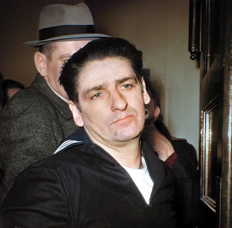 A man with slicked-back dark hair and a somber expression is escorted by another man in a suit and fedora. The background appears crowded and the setting is indoors near a doorway.