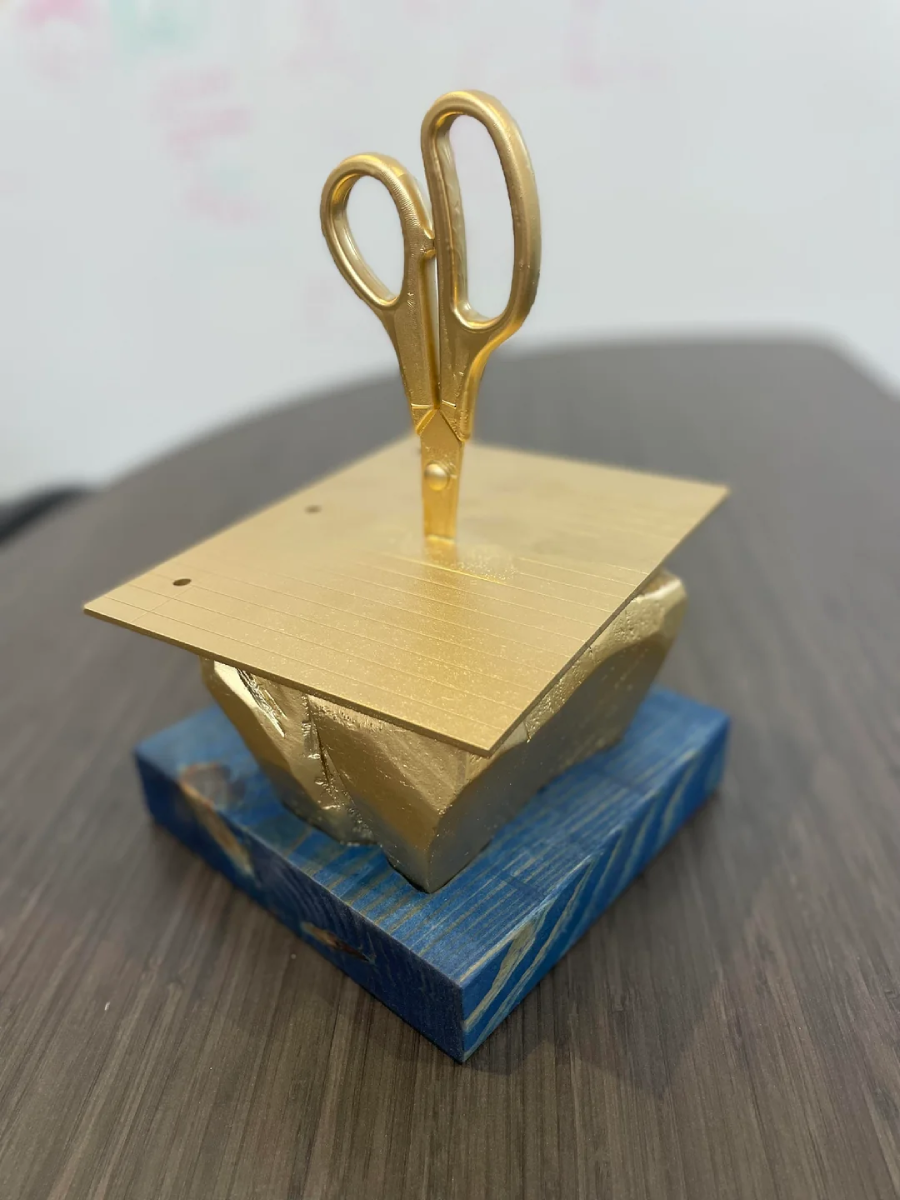 A gold-painted pair of scissors is stuck upright through a gold-painted notepad, both resting on a blue, textured base. The sculpture sits on a dark wood surface.