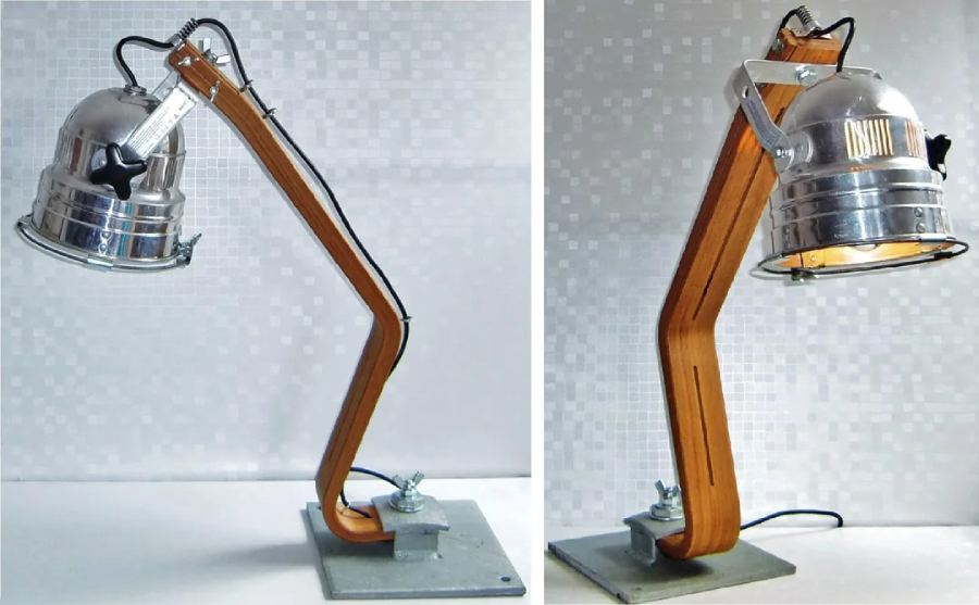 A modern desk lamp with a bent wooden arm, a metal base, and a shiny metal lampshade. The lamp has an industrial style, shown from two angles against a light, tiled background.