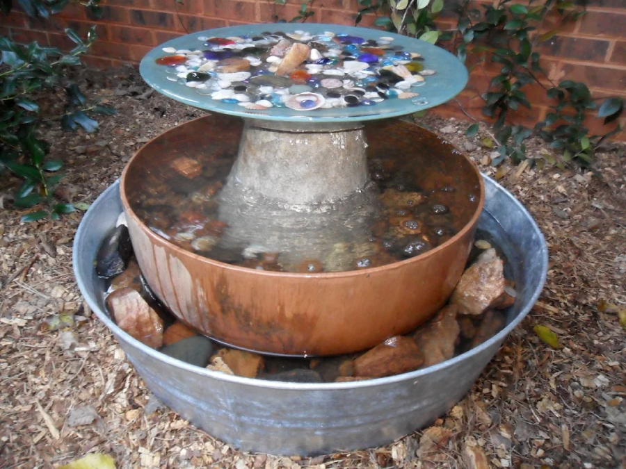 A decorative outdoor water fountain made of a copper basin in a metal tub, topped with a round glass disc embedded with colorful stones and shells. The fountain is surrounded by rocks and plants against a brick wall.