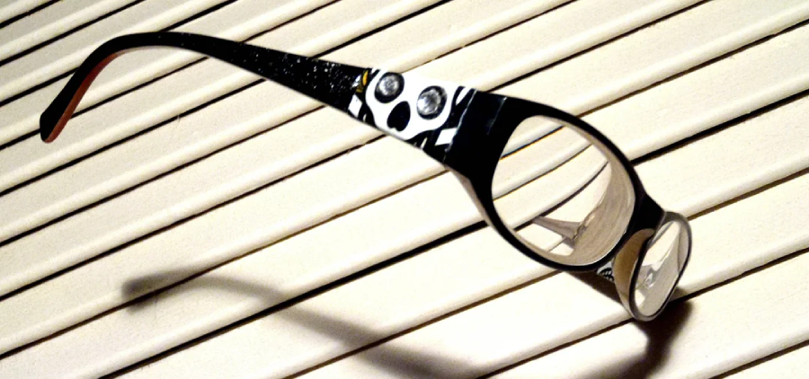 Black eyeglasses with a white skull design on the frame rest on a cream-colored slatted surface, casting a distinct shadow.