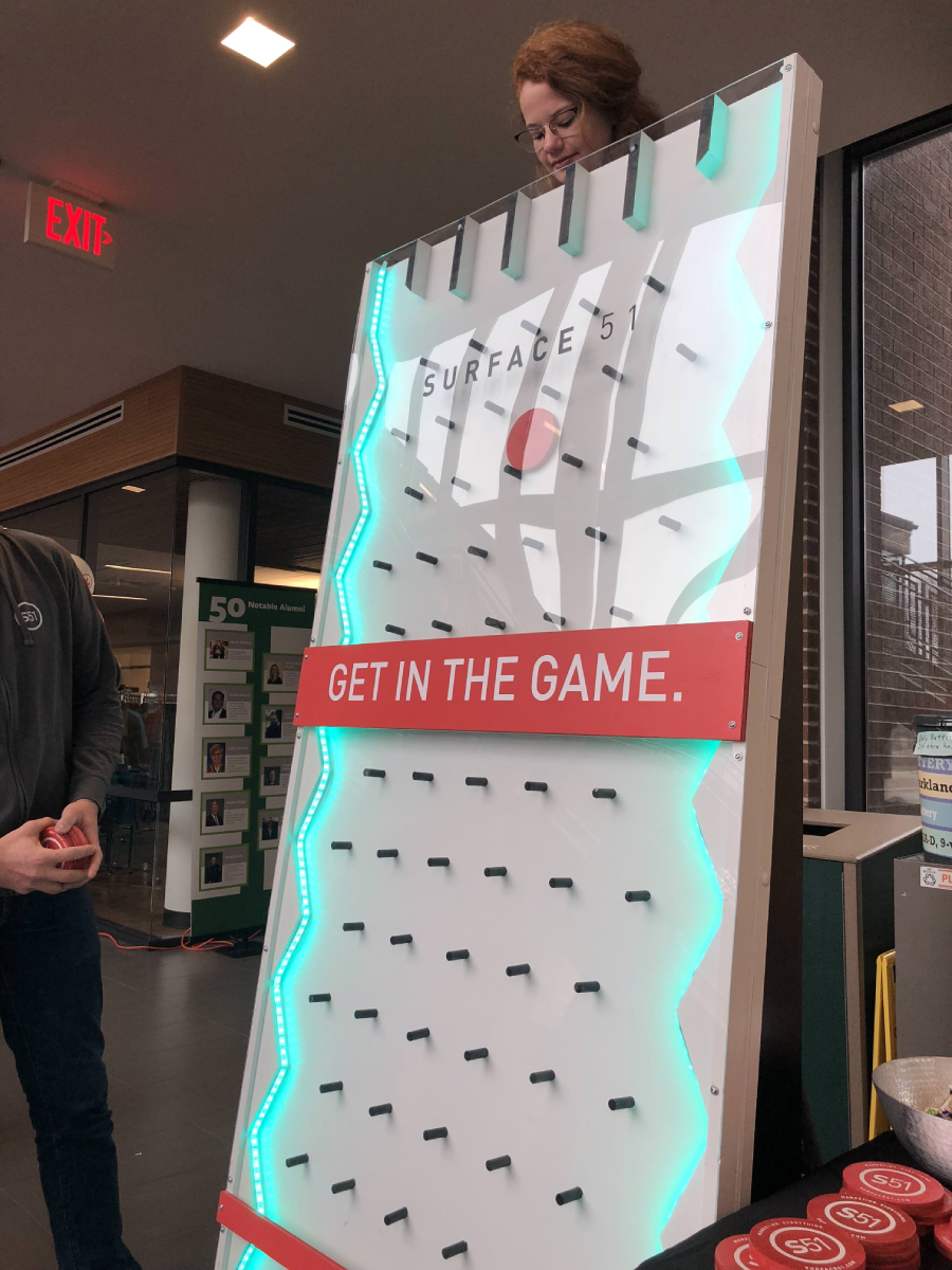 A woman drops a puck into a tall, vertical Plinko board labeled “SURFACE 51” and “GET IN THE GAME.” The board has glowing lights, and a person stands nearby with a hand visible. Indoors, with windows and photos in the background.
