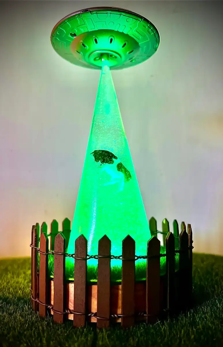A model UFO emits a green light beam, appearing to abduct a small cow figure inside a fenced grassy area, creating a playful extraterrestrial scene.