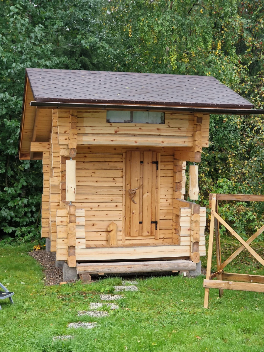 A small wooden log cabin with a dark brown roof stands on a grassy lawn, surrounded by green trees. There is a simple porch, a closed door, and a pathway of stepping stones leading to the entrance.