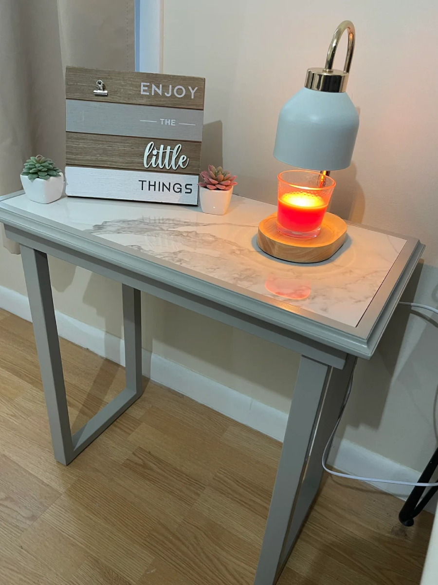 A small white table with a marble top holds a lit orange candle, a white lamp with gold accents, two small potted succulents, and a wooden sign that reads "Enjoy the little things.