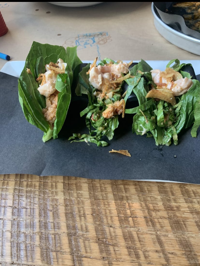 Three lettuce wraps filled with mixed greens, shredded chicken, shrimp, and crispy toppings are arranged on a black napkin over a wooden table.