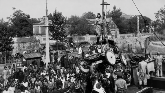 A crowd watches as a car is lifted by a crane, with a woman standing on top of the vehicle. Trees and buildings are visible in the background. The scene appears historical and draws significant public attention. Model-T