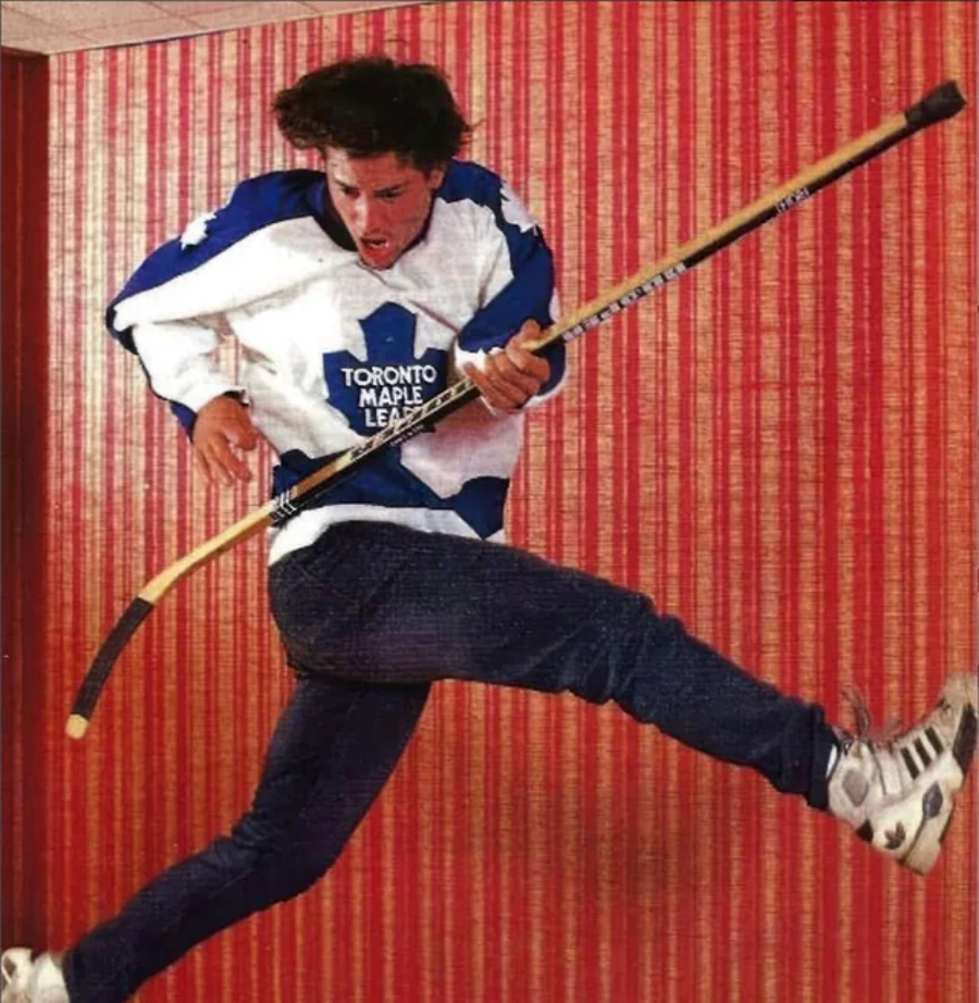 A person in a Toronto Maple Leafs hockey jersey and skates jumps mid-air against a red striped wall, holding a hockey stick horizontally across their body.
