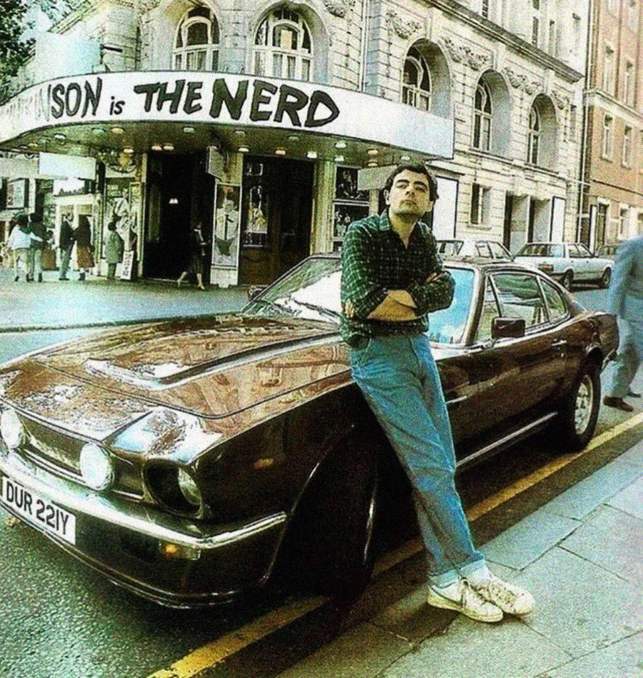 A man in casual clothes leans against a vintage dark sports car parked on a city street. Behind him is a theater with a marquee reading “SON is THE NERD.” Other cars and pedestrians are visible.