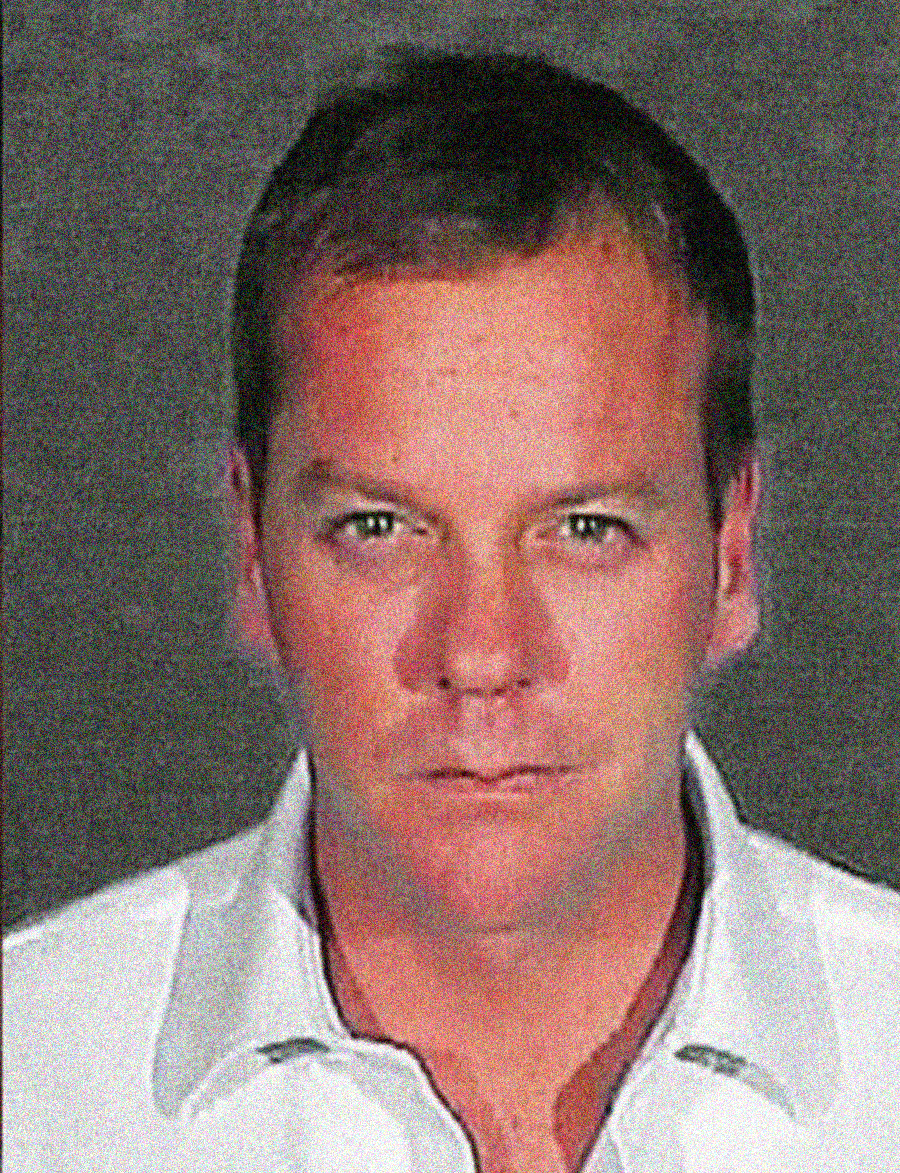 A man with short hair and a serious expression looks directly at the camera. He is wearing a light-colored collared shirt, and the background is plain and dark.
