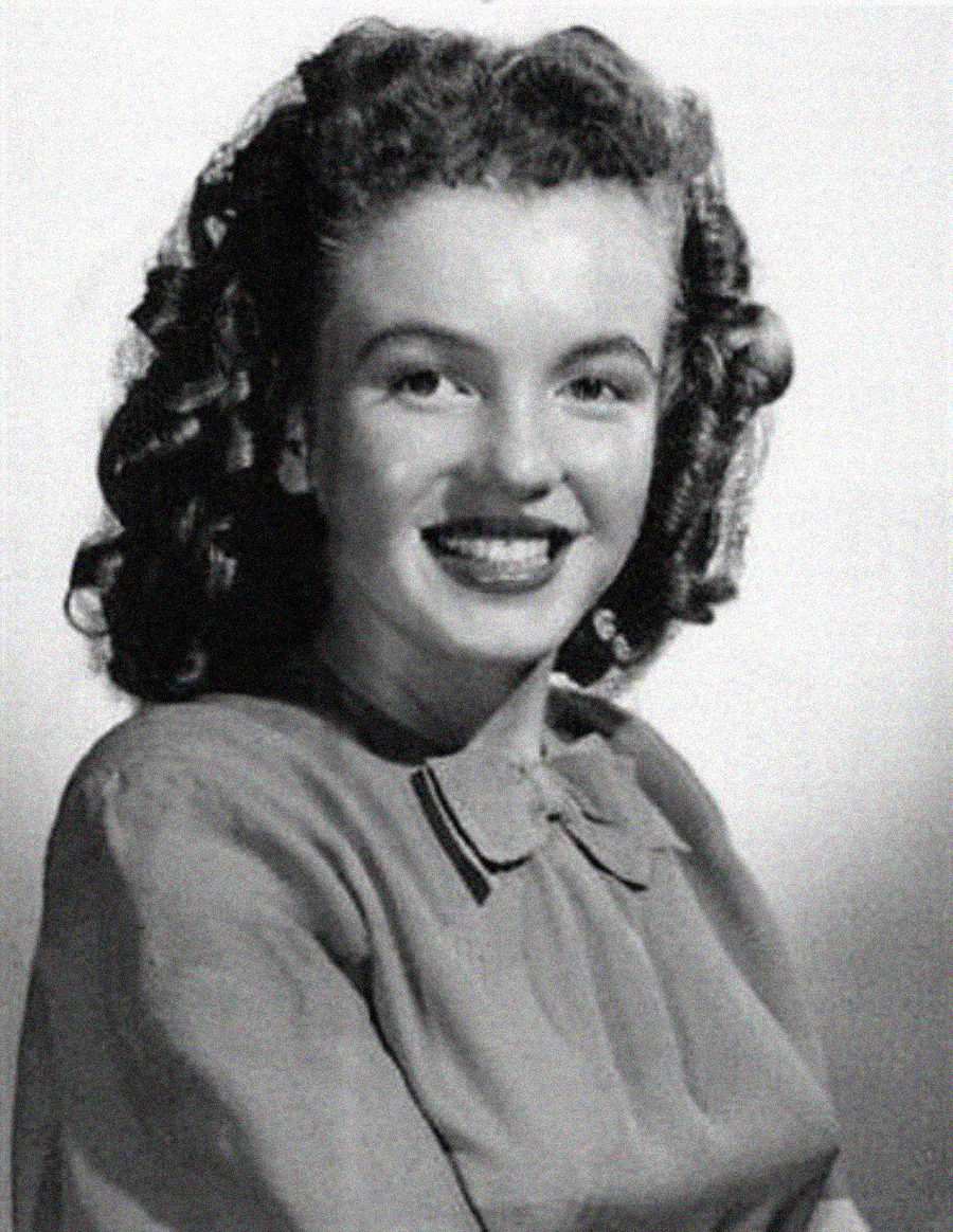 A young woman with curly hair and a wide smile poses for a black-and-white portrait. She is wearing a blouse with a small bow at the collar and is seated, looking slightly to the side.