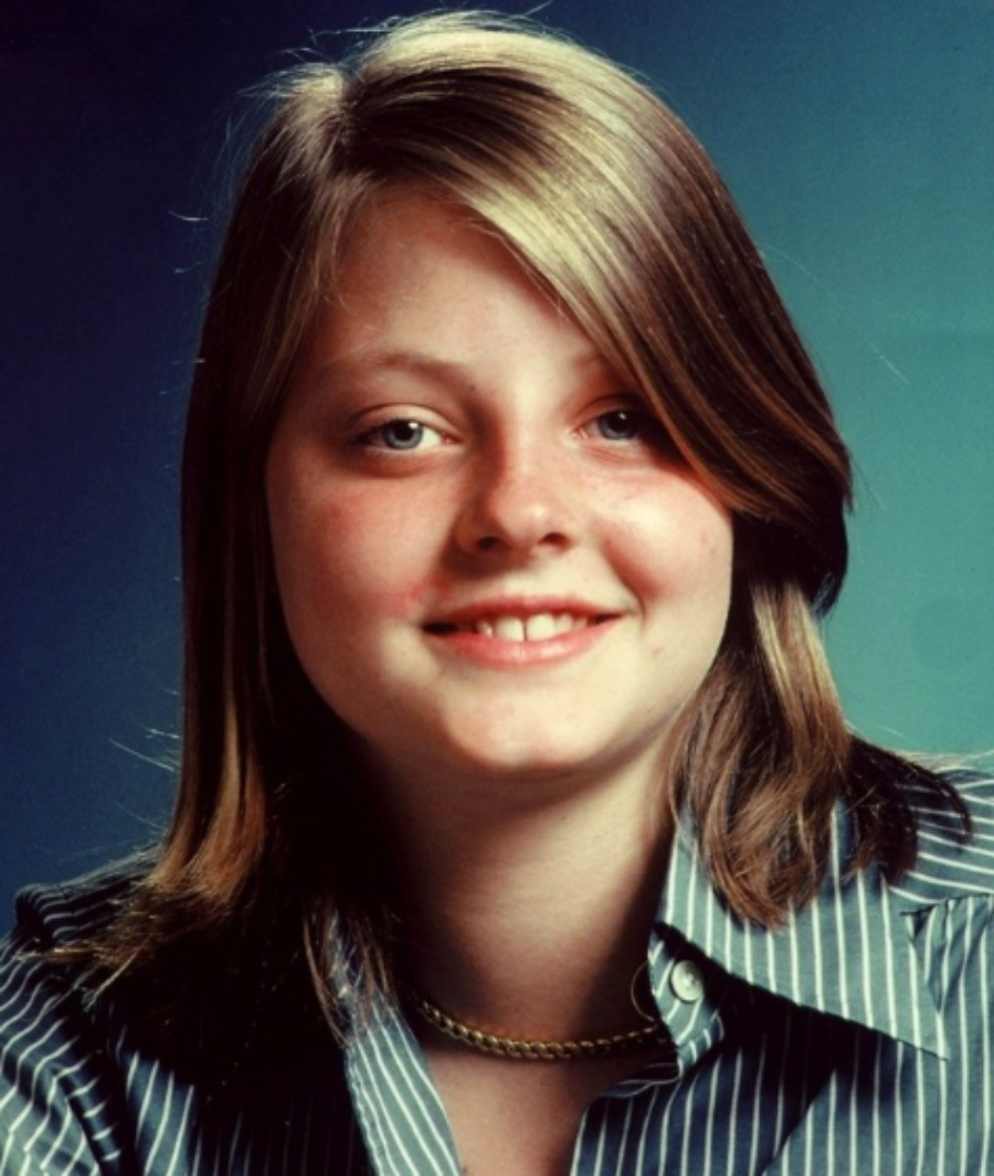 A young person with straight, shoulder-length light brown hair smiles softly while wearing a blue striped shirt and a gold chain necklace against a blue background.