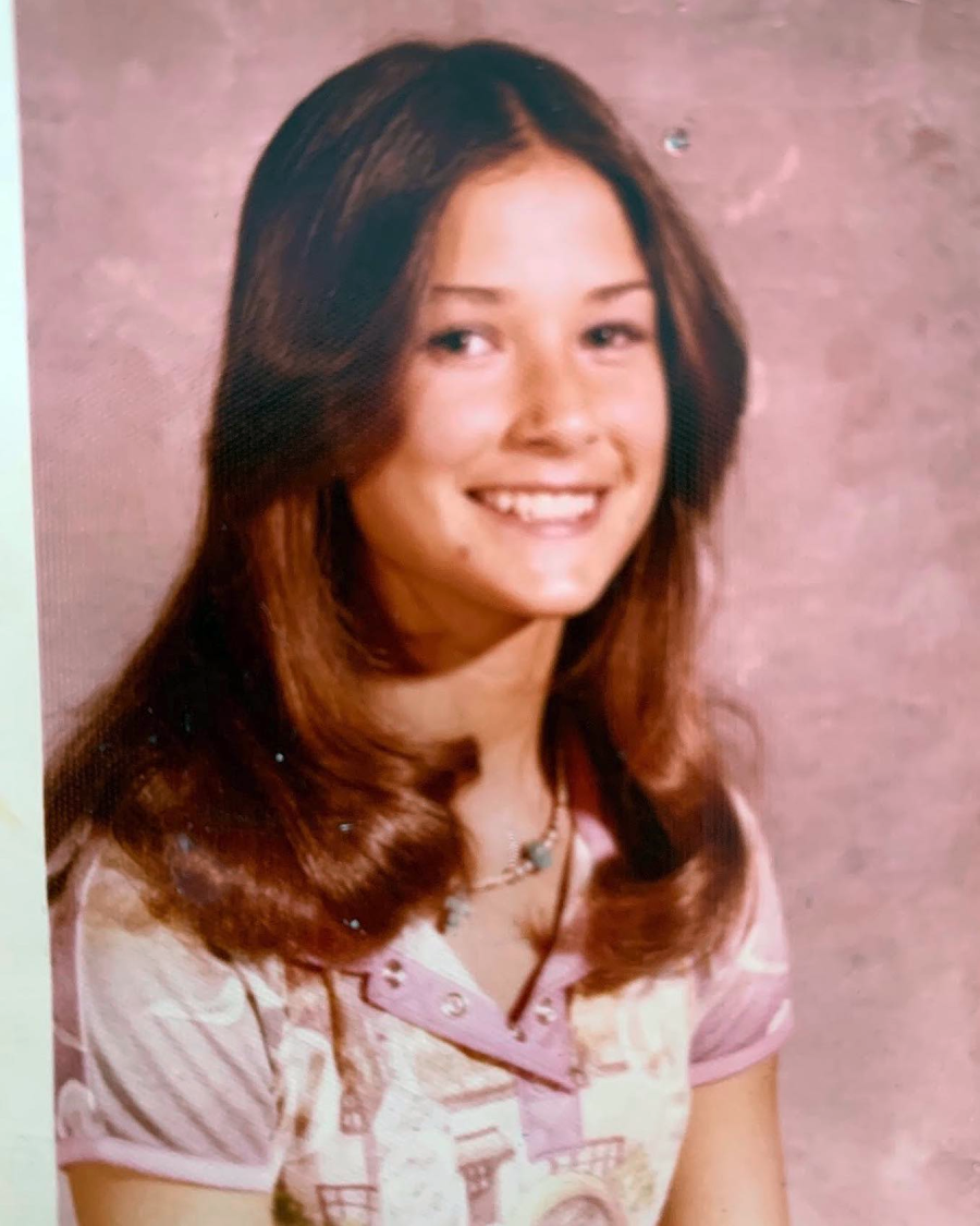 A teenage girl with long, straight brown hair and a side part smiles at the camera. She is wearing a patterned short-sleeve shirt and a necklace. The background is pink and plain, suggesting a school portrait.