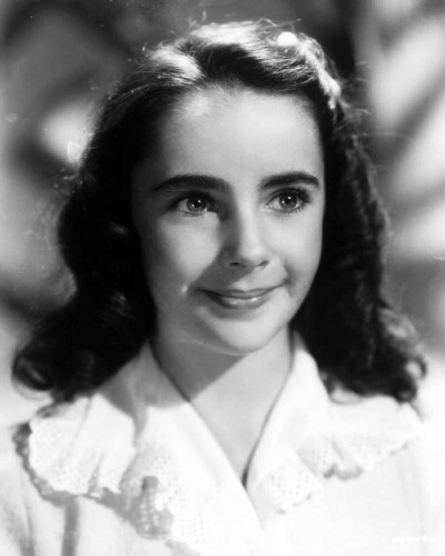 A young girl with dark wavy hair smiles softly, wearing a white blouse with a lace collar, in a black and white portrait.