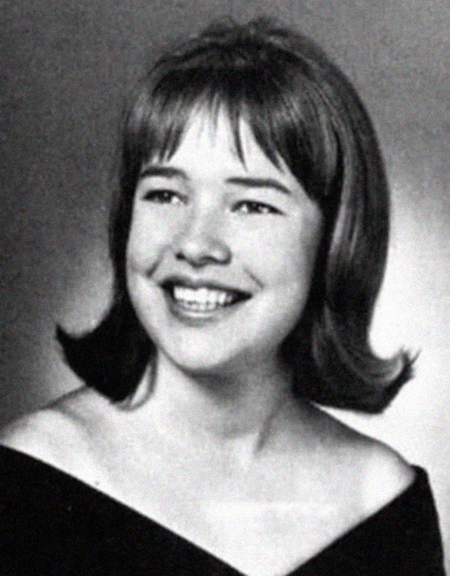 Black-and-white portrait of a young woman with chin-length hair and bangs, smiling broadly. She is wearing an off-the-shoulder dark top, and the background is plain and light-toned.
