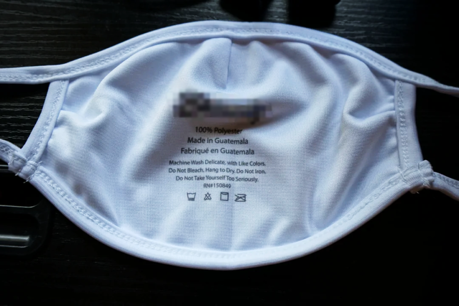 A white cloth face mask is laid flat with its inner side facing up, displaying printed care instructions and manufacturing information in black text. The mask has ear loops and is on a dark surface.