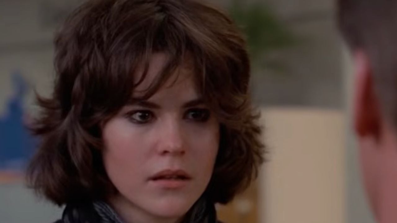 A young woman with shaggy brown hair and dark eye makeup looks ahead with a serious, slightly anxious expression. An out-of-focus person faces her in the foreground. Indoor setting with muted background colors.