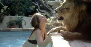 A woman in a bikini kneels at the edge of a pool, playfully facing a lion that is spraying water from its mouth toward her. Lush greenery and stone structures are visible in the background.