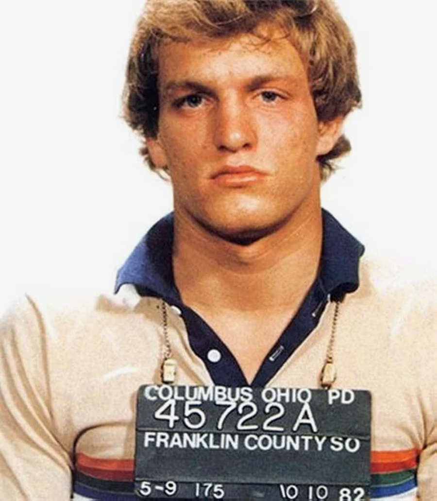 A young man with blond hair wearing a collared shirt holds a mugshot sign reading "Columbus Ohio PD, Franklin County, 10/10/82." He stands against a plain background, looking straight at the camera.