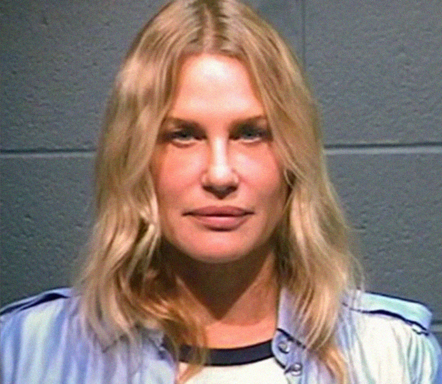 A woman with long blonde hair and a neutral expression stands against a gray cinderblock wall, wearing a light blue shirt over a white top.
