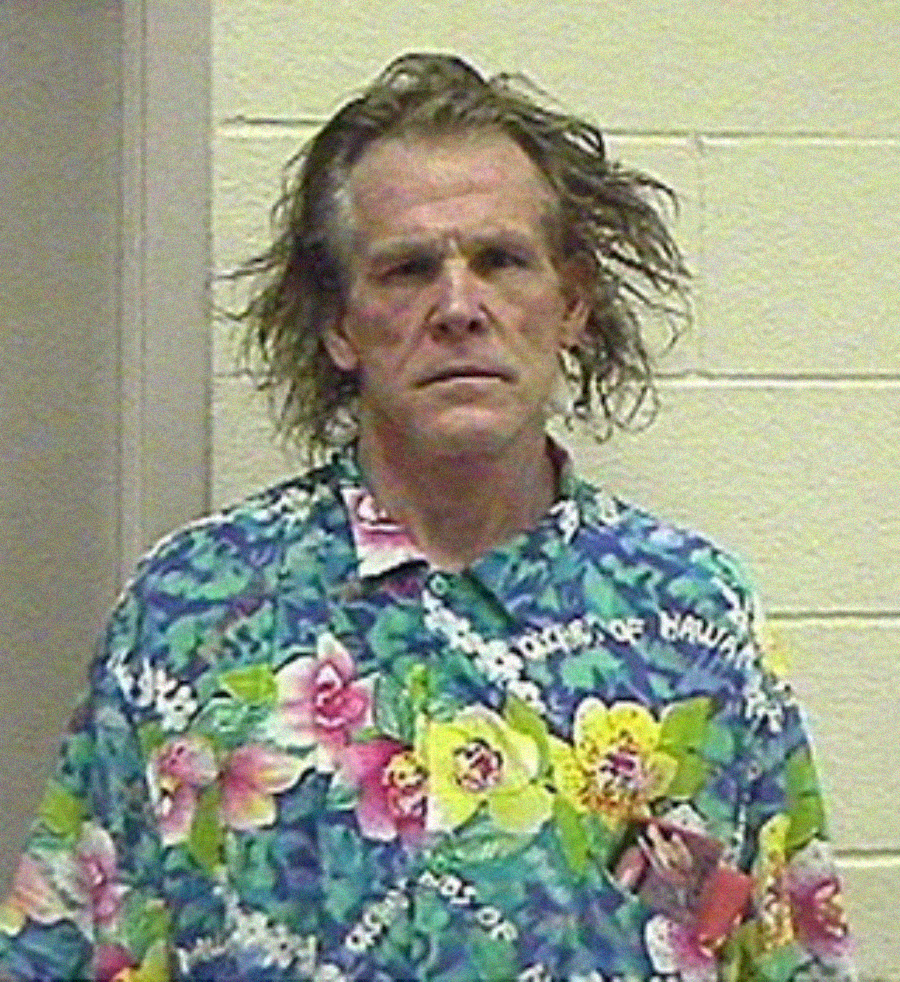 A man with messy hair wearing a brightly colored floral shirt stands against a beige wall, looking directly at the camera with a neutral expression.