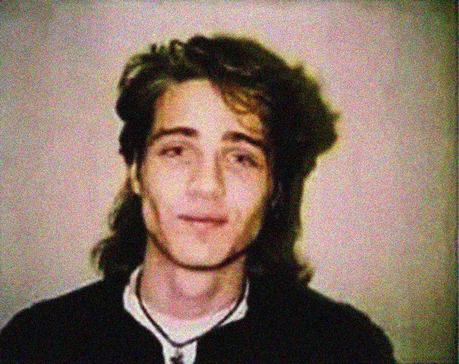 A young person with wavy, medium-length hair and a slight smile, wearing a dark shirt and a necklace, poses against a plain background in a grainy, vintage-style photo.