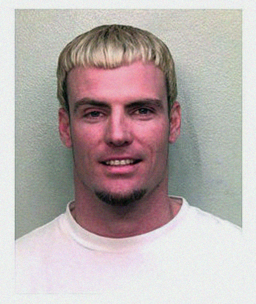 A man with short, light blond hair and a trimmed goatee smiles slightly at the camera, wearing a plain white t-shirt against a light gray background.