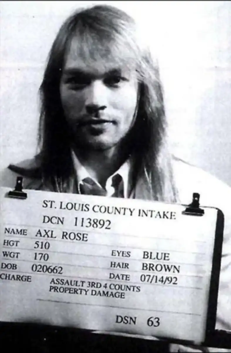Black-and-white mugshot of a man with long, light brown hair holding a sign that reads his name (Axl Rose), physical details, charges, and the date 07/14/92 at St. Louis County Intake.