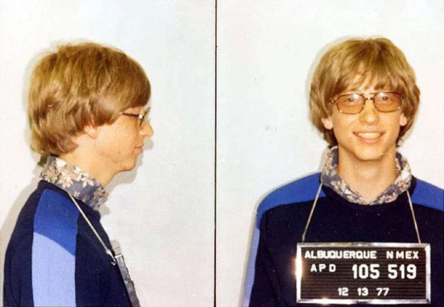 A young man with blond hair and glasses is shown in both front and side profile mugshots, smiling, wearing a blue jacket and floral shirt. The sign reads "Albuquerque N. Mex 105519, APD, 12-13-77.